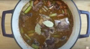 Beef stew in a pot with carrots, celery, and bay leaves simmering in rich broth on a kitchen counter.